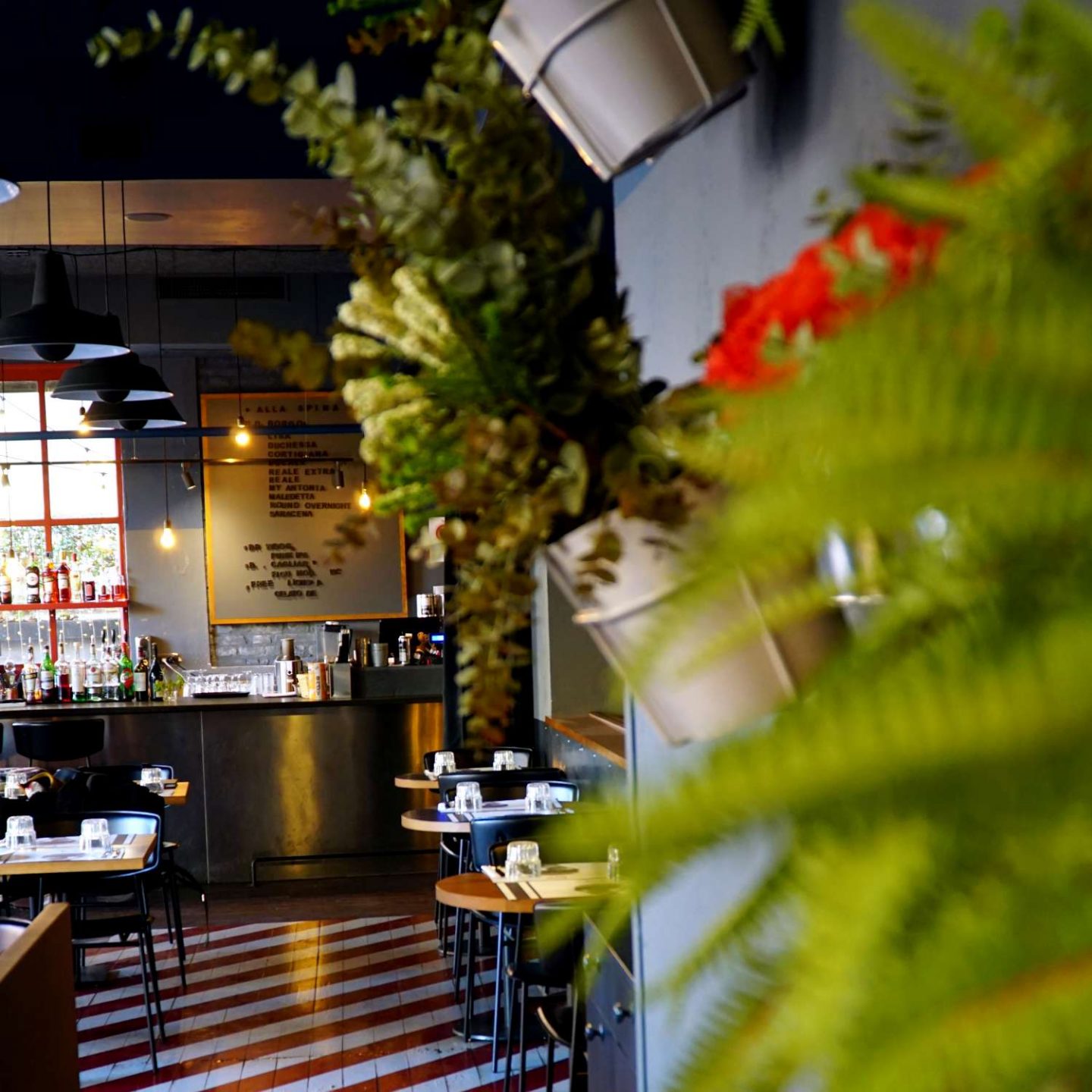 sala ristorante con tavoli di legno, pavimento a righe bianche e rosse, piante e fiori sulla parete destra, In fondo bancone con drink.