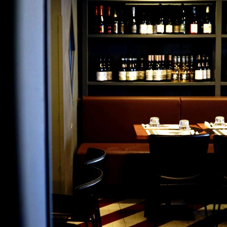 angolo della sala ristorante con tavoli in legno, tovagliette di carta e parete con bottiglie di vino e birra